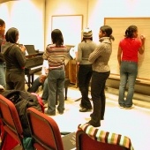 A group of students stand inside a rehearsal room talking while one writes on a chalkboard that has music bars