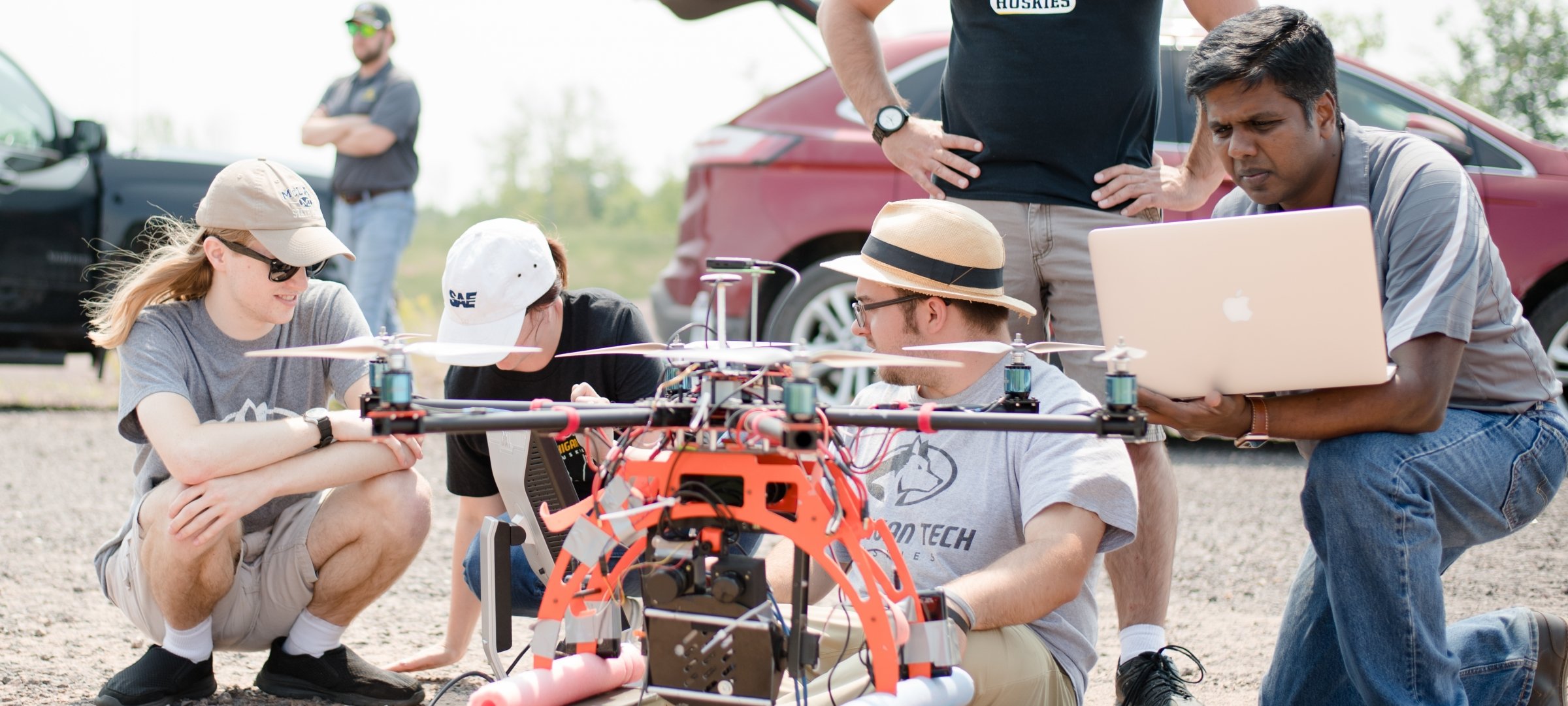 Four students outside looking at a drone in the foreground with another student behind them, hands on hips, and a man in the background near a car and truck with his arms folded.