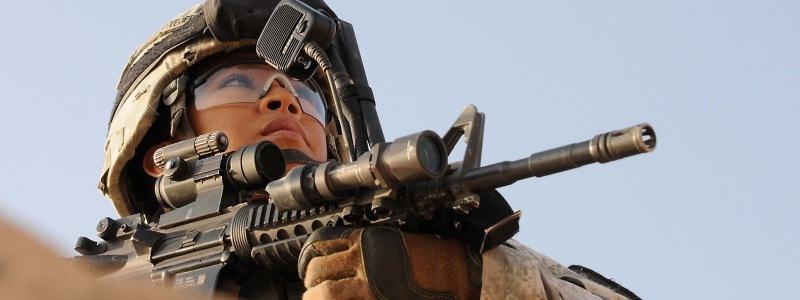 Female office, in full combat gear with riffel pointed, looks into the distance.