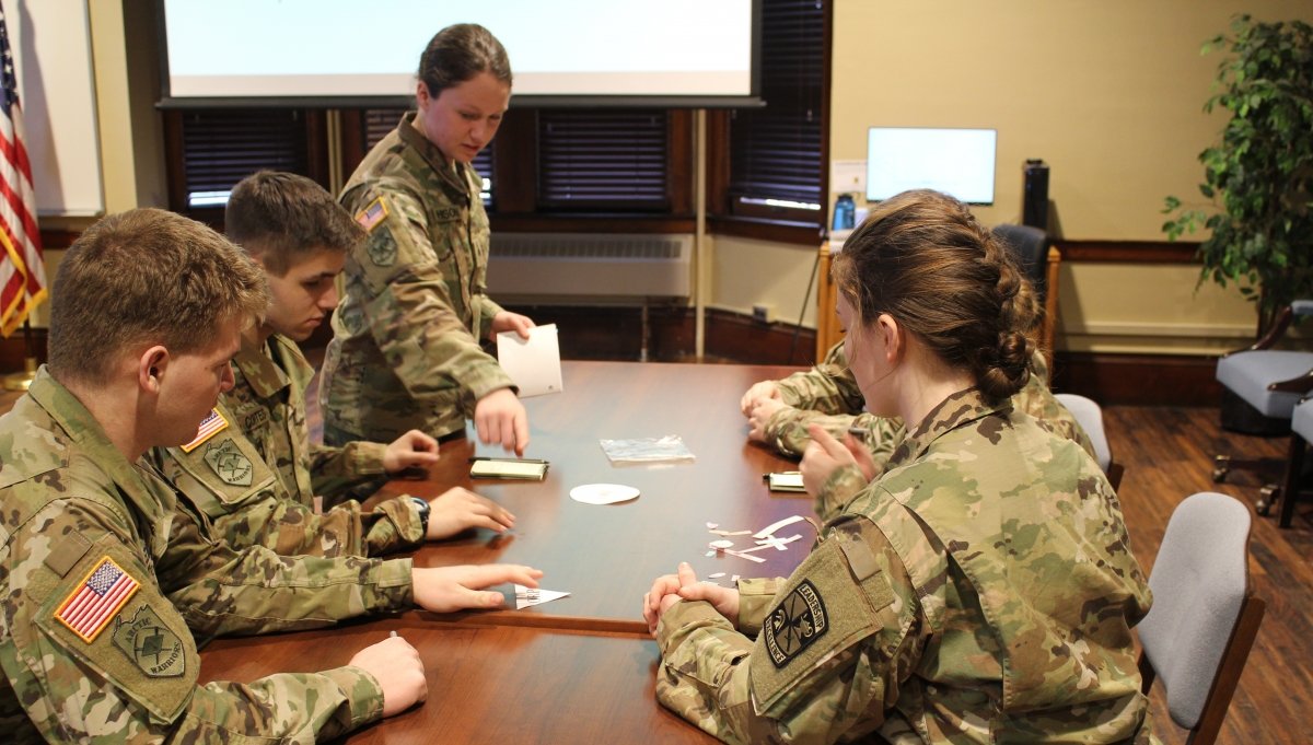 five Students around a table, wearing fatigues, briefing a mission plan. 