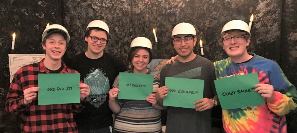 Students in an escape room at Michigan Tech holding up signs that say "We escaped" and "tenacity"