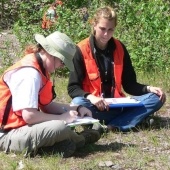 Two students with orange vests sitting outside with notebooks.
