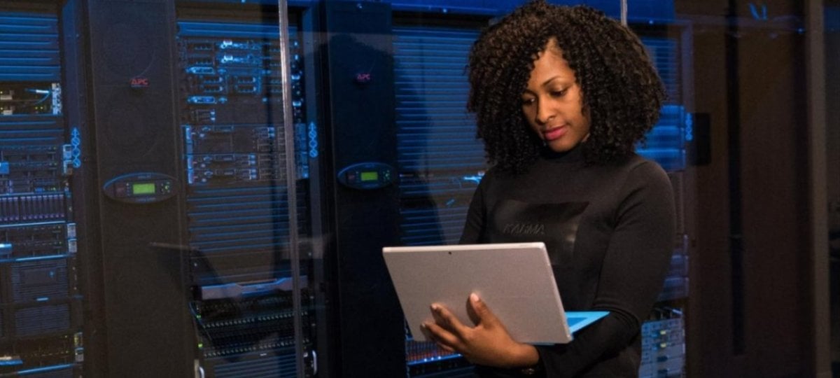 Female, African American computer engineer looking at a laptop while standing next to a mainframe computer.