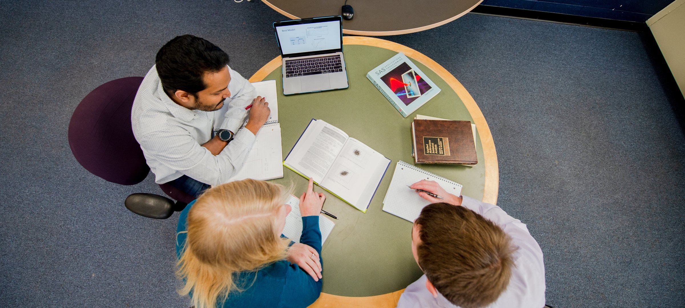 Three people looking at textbook and laptop together at a table, aerial view
