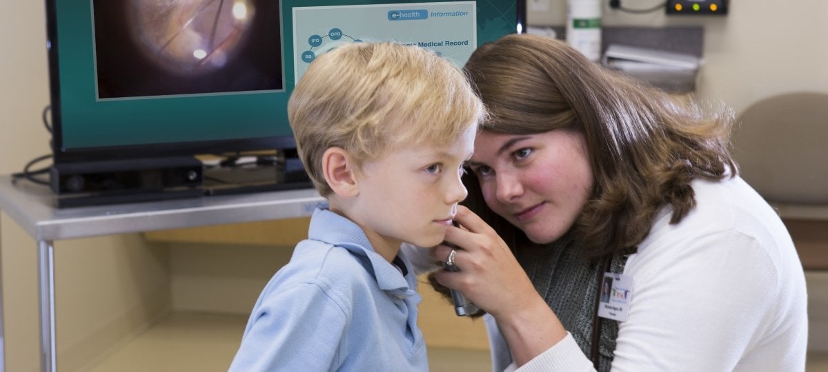 Doctor looking in the ear of a child with a computer display of patient details in the background.