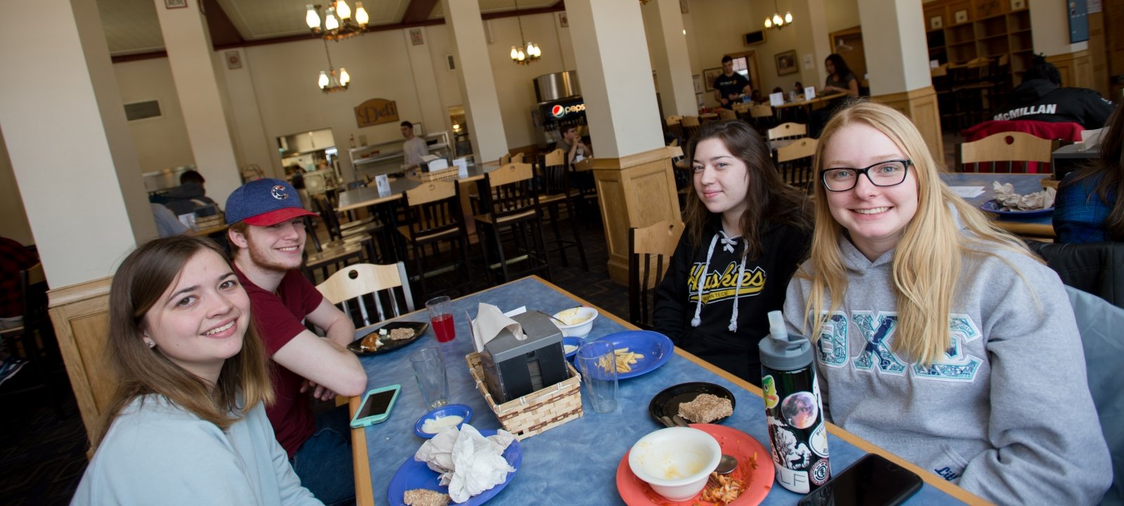 Four MTU students sit in the DHH Dining Hall with a meal, smiling at camera