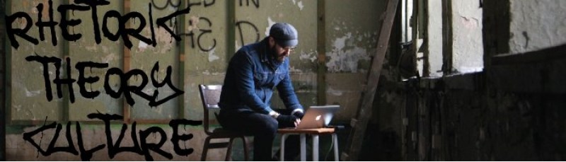 Rhetoric Theory Culture graphically overlaid on a photograph of a man sitting in a chair with a laptop on a small table in an abandoned large room