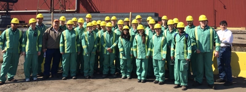 A large group of students, wearing coveralls and hard hats, pose for a photo after an industry tour. 