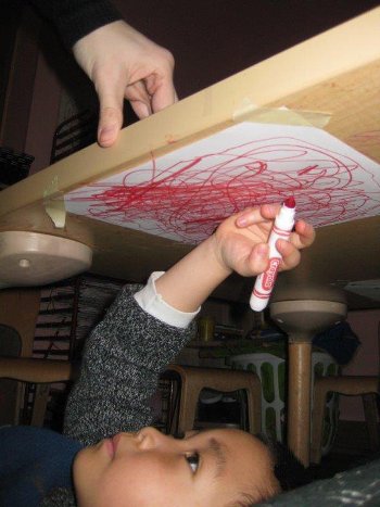 Child laying under a table coloring on a piece of paper taped there.