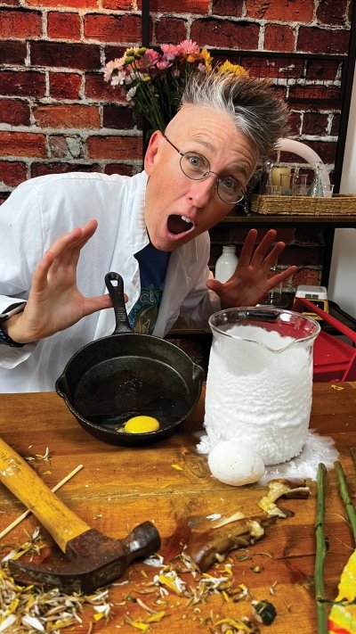 Chemical Kim looking shocked behind a table with an egg in a frying pan, a large beaker with white foam, and a hammer.