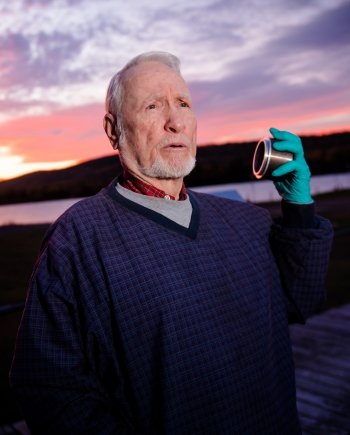 A man with short white hair and trimmed beard holds a piece of metal equipment in his gloved hand with a sunrise behind him