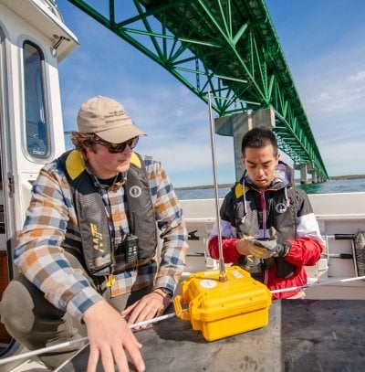 Two people on a boat under the Mackinac Bridge setting up equipment.