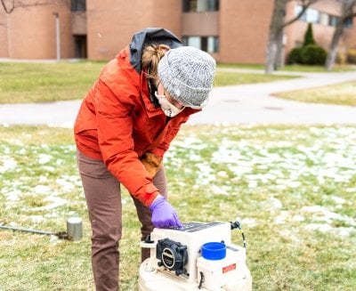 A person wearing gloves and a face mask uses an instrument outside near a waste water access point.