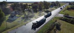 Ten trucks lay new pavement on a road through flat farmland in an aerial shot.