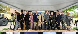 Campus leaders on a platform stage stand with black and gold shovels turning dirt during a groundbreaking ceremony.