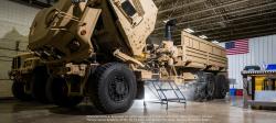 Two large tan tactical vehicles stand parked in a large warehouse structure, with the cab of the front vehicle folded up to show internal electrical components.