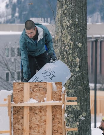 A male student holding a blue bucket standing on top of snow in a wooden form