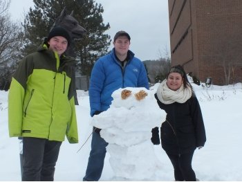 Two young men and one young female college student stand behind a snowman with dried flower eyes with a Husky statue in the background