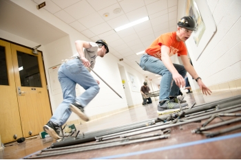Two young men wearing hard hats run metal pieces to two students building a steel bridge in the hall