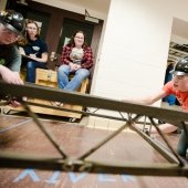 a female and male student wearing hard hats assemble a steel bridge in the hall while two female college students look on