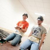 two young men with pieces of steel in a hall wearing hard hats and carpenters belts smiling