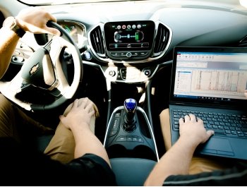 Within the cab of a car, two people sit. One holds the steering wheel and the other runs a laptop.