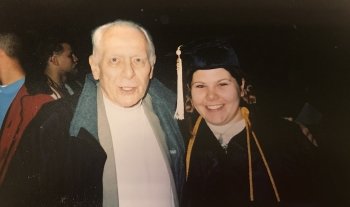 An older man and a young girl with a graduate cap on at a graduation ceremony with a dark background, black gown and gold tassel