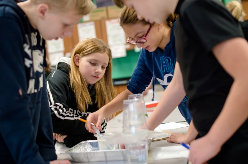 four children work on a plastic tube and water experiment in a classroom with green walls