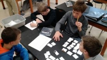 four young boys at black table with white pieces of paper on it and a see-through plastic container holding pencils in a science classroom looking at a science and engineering problem to solve