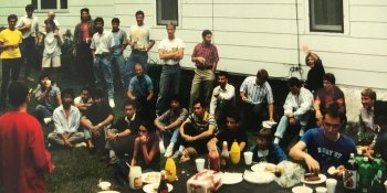 a group of young men and women by a picnic table sitting and standing on the grass with a white sided house in the background eating food together at an international cookout
