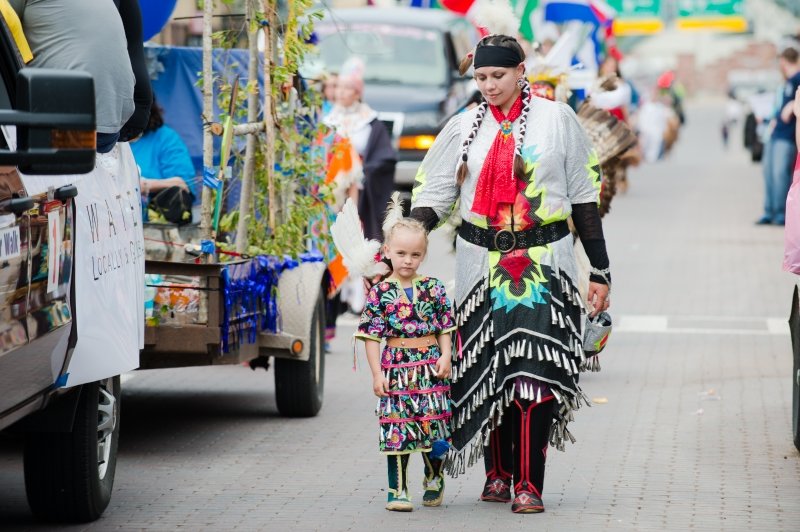 A mother and her little daughter walk in native american regalia walk next to a float with branches on it on a downtown street outside in a parade