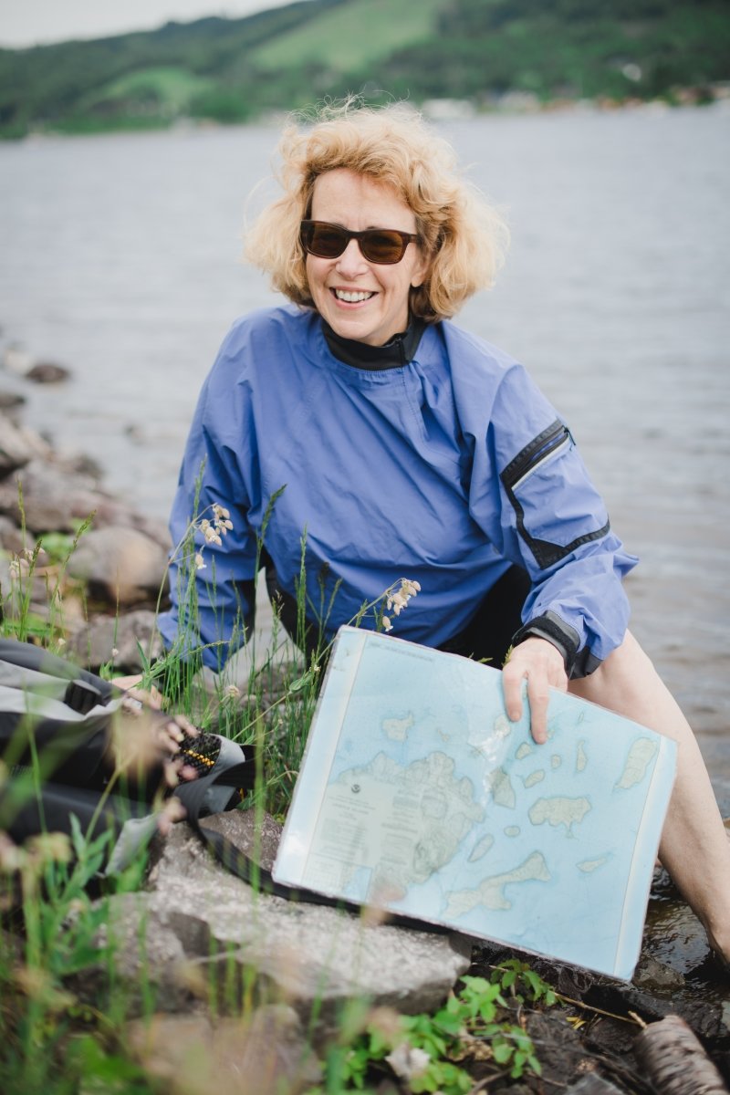 A woman sitting on rocks on the shore of a canal/river holding a map of lake superior and a waterproof bag, wearing sunglasses and smiing
