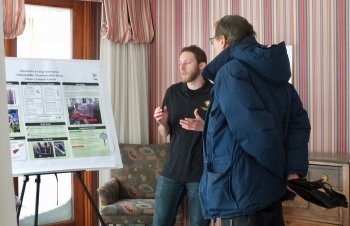 A young man talks to an older man showing him a poster of a composting project in a house with pink striped wallpaper with snow outside.