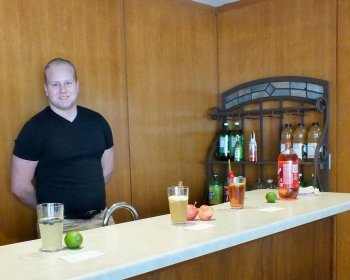A young man stands behind a wooden bar with wood paneling in the background with soft drinks, glasses and fruit on the bar.
