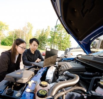 two people working on a laptop under the hood of a car