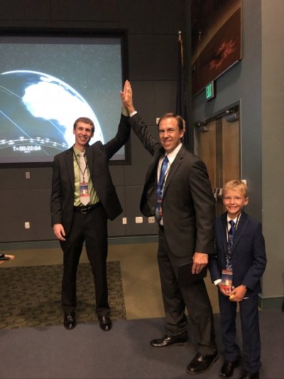 two men is suits and ties do a high five with a planetary screen and an exit sign behind them while a young boy in a suit and tie smiles in front of them