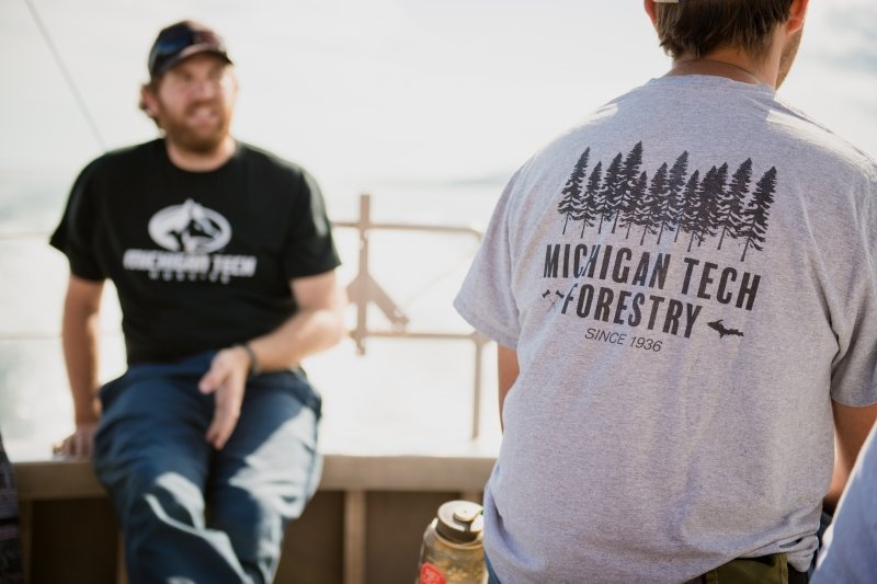 A man with a beard and cap and a Michigan Tech t-shirt faces the camera and a man with a Michigan Tech School of Forestry shirt shows the back of his shirt to the camera with water spray on a boat in the background.
