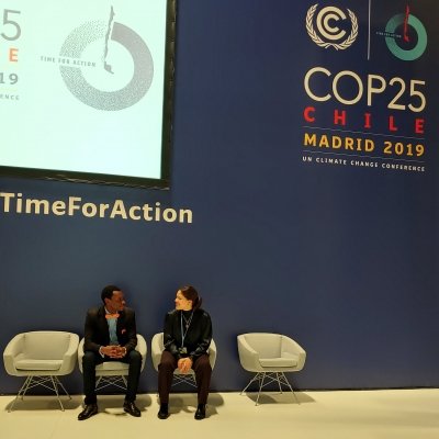 A man and a woman, both seated, looked at each other while smiling. They sit underneath a banner that reads &quot;COP25 Chile Madrid 2019 UN Climate Change Conference&quot;.
