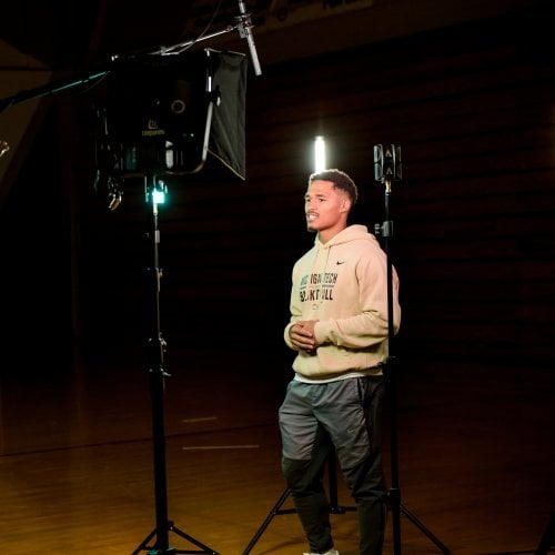 A young man wearing a Michigan Tech football shirt stands under studio lights in a wood gym.