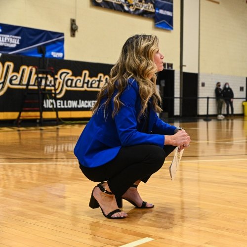 A woman squats on a gym floor with a blue hornets sign behind her coaching a volleyball game.