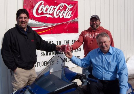Gary and Rob Scholie (standing, left to right), owners of Coca-Cola of Hancock, celebrate the Coca-Cola Foundation?s $10,000 donation to the SAE Clean Snowmobile Challenge with Challenge organizer Jay Meldrum.