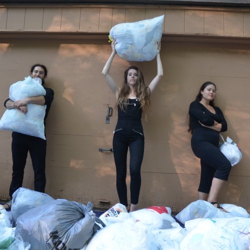A young man in center and two young women stand holding a plethora of plastic bags in front of a wooden garage wall