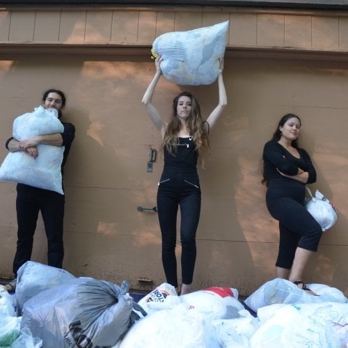 A young man in center and two young women stand holding a plethora of plastic bags in front of a wooden garage wall