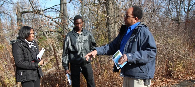 A Black educator talks with two Black High School students about natural resources careers on the sidewalk next to a late-fall forest in a STEM outreach program focused on the environment