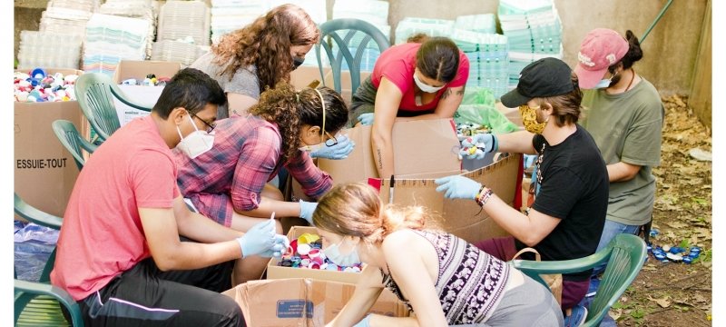 six young people in masks and gloves sort recyclables outside with a brick house and trees in the background