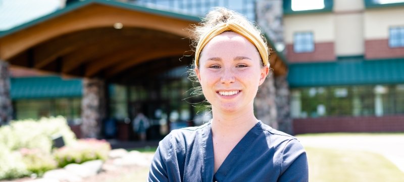 A young woman in scrubs smiles in front of a hospital outside with a blue sky in Hancock, Michigan