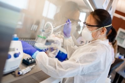 A person wearing a lab coat, face mask, and protective eyewear uses a pipette under a vent hood.