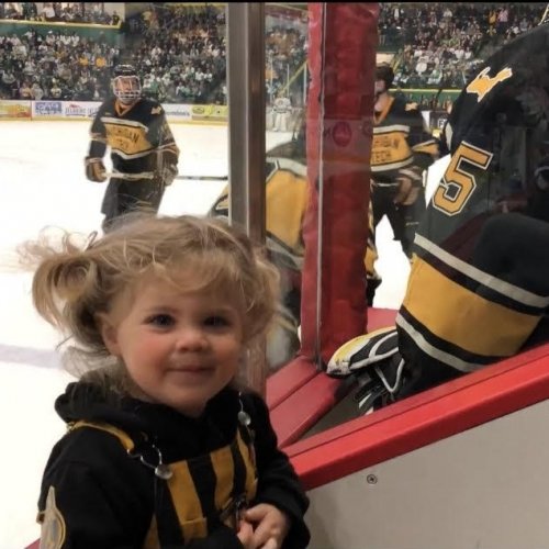 A little girl in pigtails smiles next to the Michigan Tech hockey bench with players on it with other players on the ice in the background