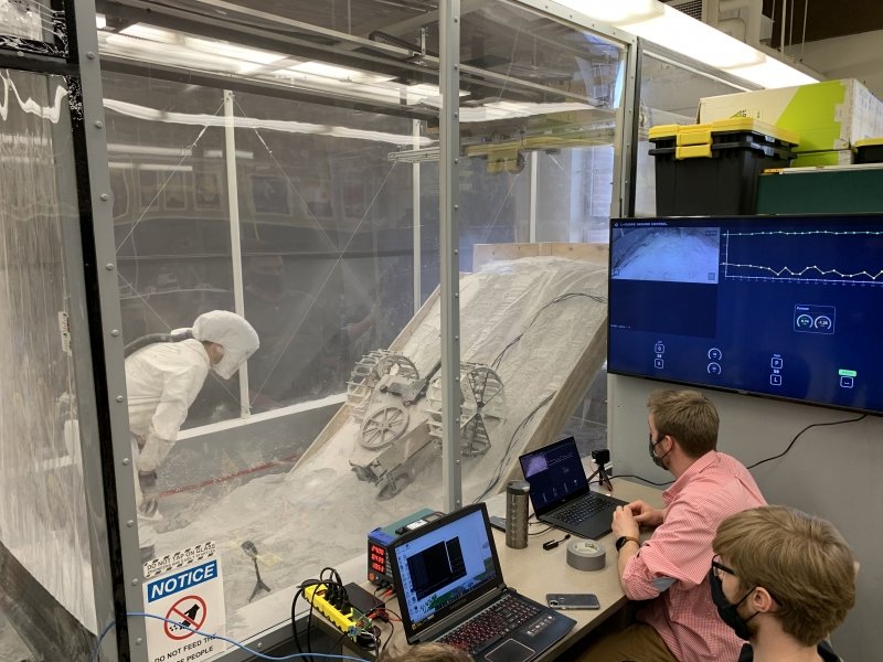 student in protective gear stands in a sealed lunar test chamber with a rover