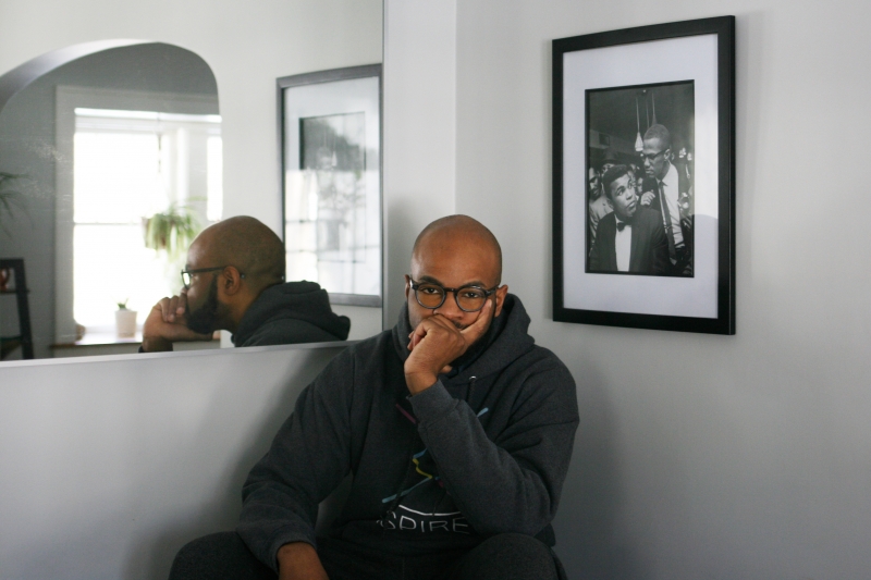 A young man with chin cupped in hand reflects in front of a portrait of Mohammed Ali and Malcom X.